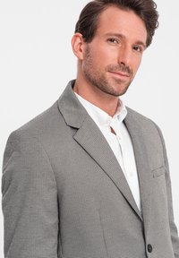 Grau karriertes Blazer mit Reverskragen, einer einzigen Brusttasche und schwarzen Knopfdetails. Getragen über einem weißen Hemd.