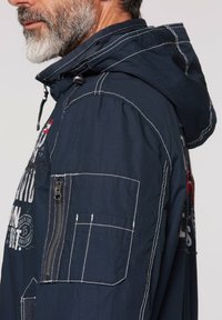 Navy hooded jacket with white stitching, zippered pocket on the arm, and graphic prints on the front and back. Lightweight fabric design.