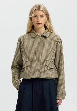 Femme aux longs cheveux blonds portant une veste beige à carreaux avec poches à rabat et un pantalon large sombre, debout devant un fond uni.