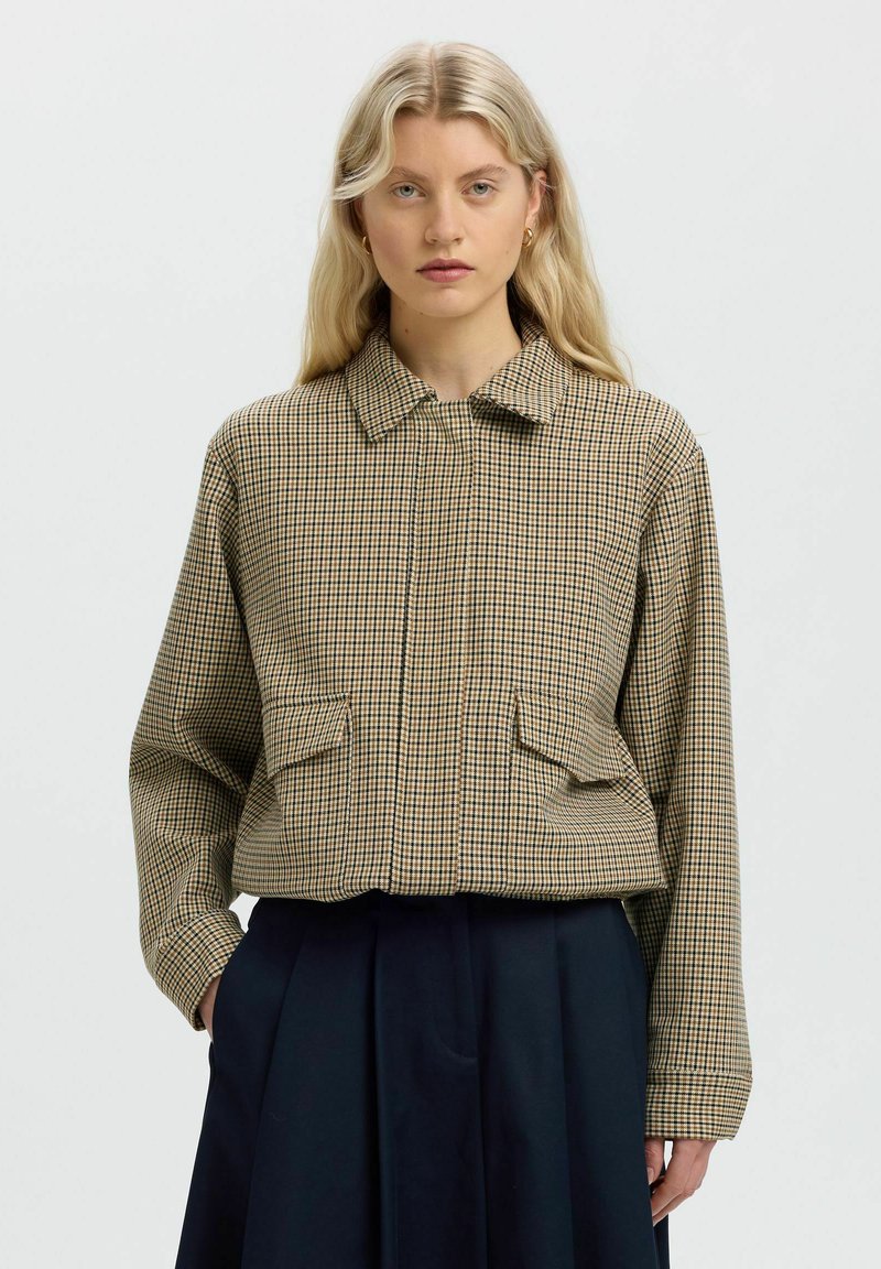 Femme aux longs cheveux blonds portant une veste beige à carreaux avec poches à rabat et un pantalon large sombre, debout devant un fond uni.