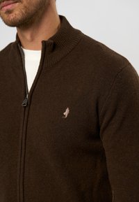 Lengvas rudas zip-up megztinis su piniotu apykakle. Yra subtilus siuvinėjimas šviesiai bežėje spalvoje ant krūtinės. Minkšta tekstūra.