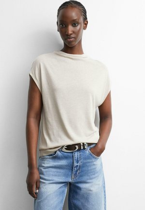 Frau mit geflochtenem Haar, die ein ärmelloses, beiges Oberteil trägt, das in hellblaue Jeans gesteckt ist, mit einer Hand in der Tasche vor weißem Hintergrund.