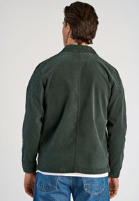 Donkergroene corduroy jas met een kraag, zichtbare naden en een ontspannen pasvorm. Gedragen over een wit shirt, in combinatie met blauwe denim jeans.