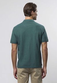 Tumši zaļa polo krekls ar īsām piedurknēm, apkakli un taisnu apakšu. Materiāls šķiet mīksts ar minimālu tekstūru un bez redzamiem rakstiem.