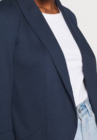 Marinblå blazer med slag, strukturerade axlar och sidofickor, matchad med en vit ribbad topp och ljusblå höga jeans.