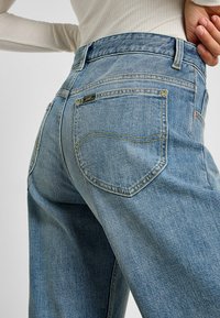 Jeans in denim azzurro con vestibilità rilassata, dotati di tasche posteriori e dettagli in cucitura gialla. Il materiale appare strutturato con un leggero lucido.
