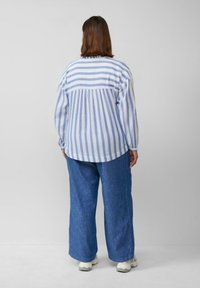 Gestreepte blauwe en witte blouse met lange mouwen, een geplooide rugpas en een relaxte pasvorm, gecombineerd met losse blauwe denim broek en witte sneakers.