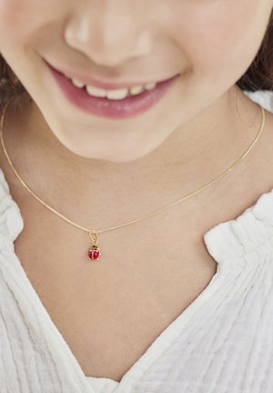Collana dorata con un pendente sferico rosso caratterizzato da una superficie texture. La catena è delicata e la camicia è in un morbido tessuto bianco.