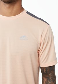 Camisetta rosa chiaro a maniche corte con tessuto testo, accenti grigi sulle spalle e un piccolo logo blu a forma di montagna sul petto.