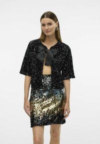 Chaqueta corta de manga corta negra con lentejuelas y un gran lazo negro en el escote, combinada con una falda brillante de lentejuelas en tonos dorados y plateados.