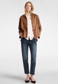 Ruskea nahkavaatetus nappikiinnityksellä, valkoinen kauluspaita, tummansiniset farkut ja ruskeat tasapohjaiset kengät. Sileä pinta, rento siluetti.