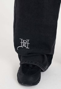Black denim pants with a loose fit, featuring white embroidered logo detail at the hem, paired with a black sneaker.