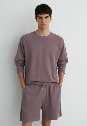 Jeune homme portant un sweat-shirt texturé mauve à manches longues et un short à cordon assorti, debout les mains dans les poches devant un mur uni.