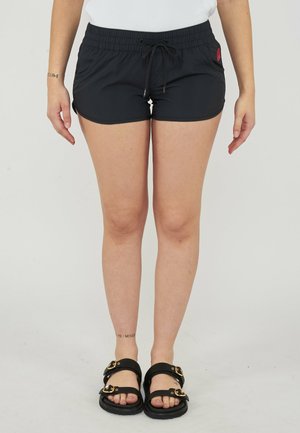 Persona che indossa pantaloncini neri con coulisse, sandali neri con fibbie dorate e una maglietta bianca, in piedi su uno sfondo chiaro e uniforme.