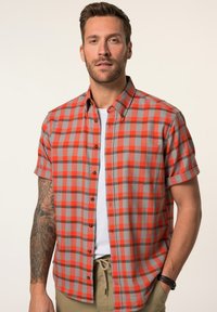 Camisa de cuadros de manga corta en rojo y gris, con cuello abotonado, bolsillo en el pecho y un patrón de cuadros en contraste. Lleva una camiseta blanca debajo.