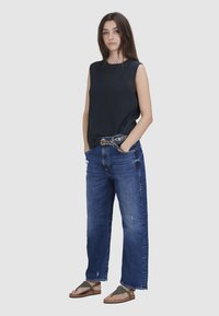 Dunkelblaues ärmelloses Oberteil aus strukturiertem Stoff, kombiniert mit locker sitzenden, verwaschenen blauen Jeans und braunen Sandalen mit gemusterten Riemen.