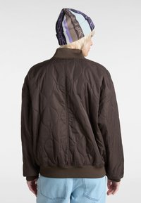 Braune gesteppte Bomberjacke mit gerippten Manschetten und Saum, die eine glatte Textur aufweist. Trägt dazu hellblaue Jeans und eine gemusterte Mütze.