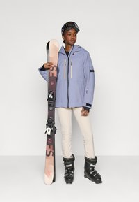 Violetti vedenpitävä hiihto takki, jossa on kaksi keltaista vetoketjua, mustat yksityiskohdat, vaaleanbeiget hiihtohousut, mustat hiihtokengät ja lumilauta siteillä.