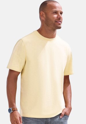 Mann trägt ein blassgelbes kurzärmeliges T-Shirt und graue Hose, hat eine Armbanduhr am linken Handgelenk und steht mit der linken Hand in der Tasche.