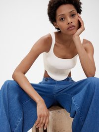 Bílý žebrovaný crop top s širokými ramínky, zkombinovaný s oversized modrými džínovými kalhotami. Přírodní dřevěný povrch pod sedící modelkou.