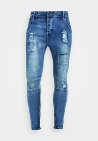 Skinny fit blue jeans with distressed patches, varying shades, and frayed accents. Made of denim with a smooth texture and standard five-pocket design.