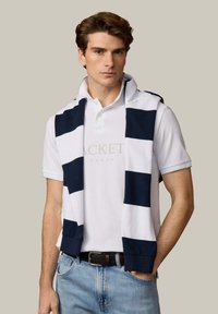 Jeune homme portant un polo blanc et un pull à rayures bleu marine et blanches posé sur les épaules, avec un jean bleu clair et une ceinture marron.