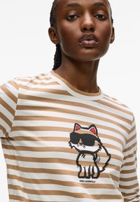 Randig beige och vit t-shirt med kortärmad design som visar en tecknad katt med solglasögon, tryckt i svart och vitt, med en logotyp under.