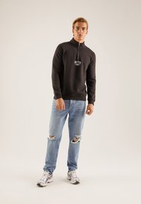 Sudadera negra con cremallera y cuello alto, con bordado de "BOSTON", combinada con unos vaqueros claros desgastados y zapatillas deportivas plateadas.