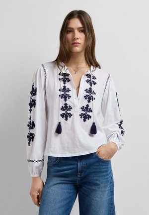 Vrouw draagt een wit blouse met lange mouwen, marineblauwe bloemenborduursels en kwastjes, gecombineerd met een blauwe spijkerbroek, staand tegen een effen achtergrond.