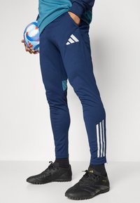Marineblå træningsbukser med hvide striber og Adidas-logo, lavet af et glat stof. Sorte sportssko med teksturerede såler. Blå fodbold holdt.