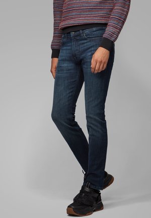 Person wearing dark blue slim-fit jeans, black sneakers with brown soles, and a multicolored striped sweater with black cuffs.