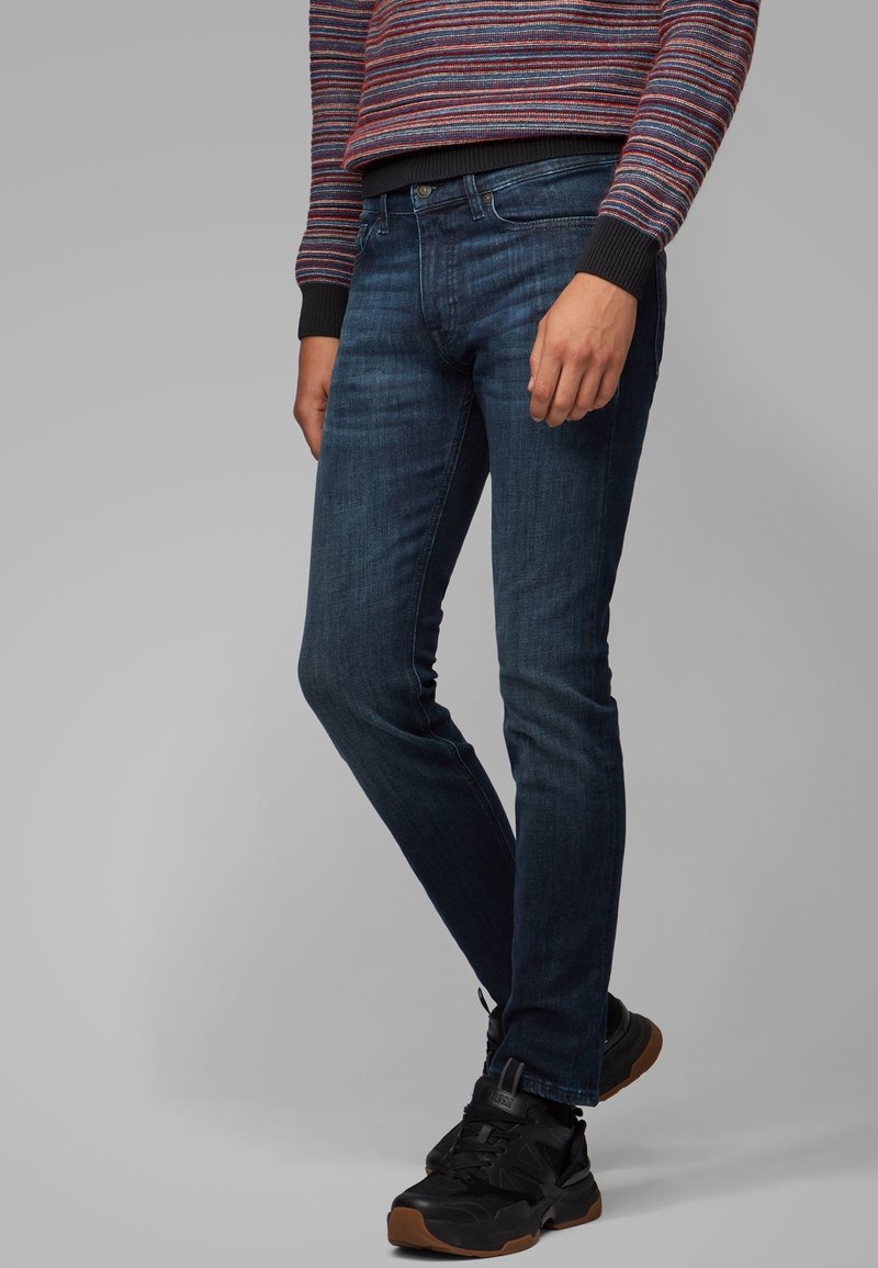 Personne portant un jean slim bleu marine, des baskets noires avec des semelles marron, et un pull rayé multicolore avec des poignets noirs.