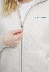 Grå hettejakke med minimalistisk design, med en sølvglidelås og merkevarenavnet "CASPARA" i grønn på venstre side.