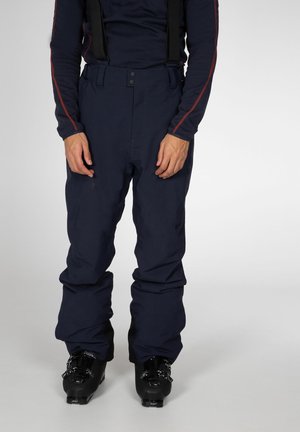 Person iført navy skibukser med seler og sorte skistøvler mod en ensfarvet grå baggrund.