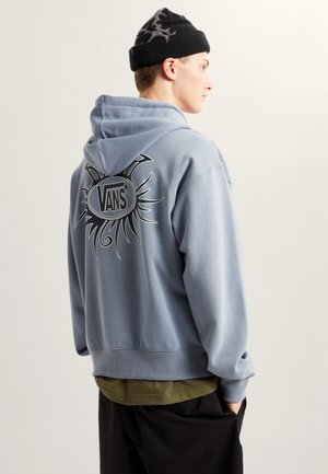 Helleblauer Kapuzenpullover mit Kapuze, auf dem Rückenteil mit einem Sonnenmotiv und dem "VANS"-Logo versehen. Getragen über einem olivgrünen Shirt und schwarzer Hose.