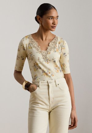 Femme portant un haut floral crème avec un col en V en dentelle et un pantalon beige taille haute, regardant sur le côté, main dans la poche, avec des bracelets et des boucles d'oreilles.