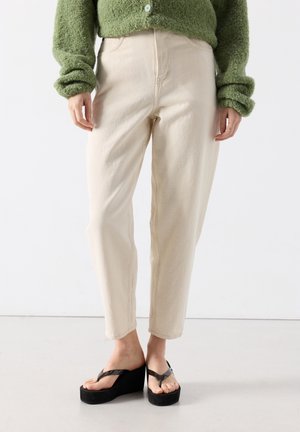 Personne portant un pantalon beige taille haute, un pull vert duveteux et des tongs plateformes noires debout sur un sol blanc.