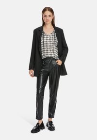 Schwarze Blazer über einer Bluse im schwarz-weißen Hahnentrittmuster, kombiniert mit glänzenden schwarzen Lederhosen und klobigen schwarzen Schuhen.