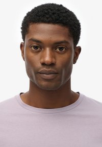 T-shirt lavande à col rond porté par une personne avec des cheveux courts et bouclés noirs et une peau lisse. Les caractéristiques notables incluent des yeux sombres expressifs et des contours faciaux subtils.