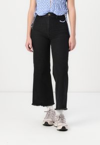 Jeans larges noirs en denim avec un ourlet effiloché et taille haute. Associés à des chaussures de sport rose clair et un haut à motifs bleus.