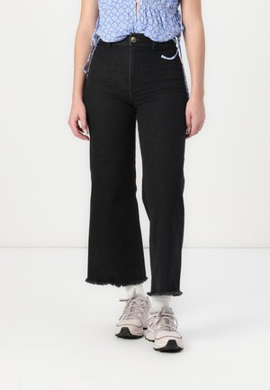 Jeans larges noirs en denim avec un ourlet effiloché et taille haute. Associés à des chaussures de sport rose clair et un haut à motifs bleus.