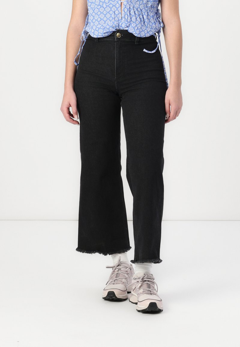 Svarta vida denimjeans med fransad nederkant och hög midja. Kombineras med ljusrosa träningsskor och en blåmönstrad topp.