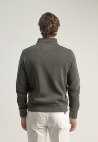 Sudadera gris de canalé con cuello alto, que cuenta con un yugo trasero y puños elásticos, combinada con pantalones de color claro.