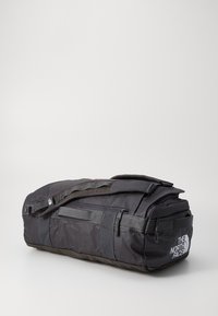 Black duffel bag with a curved shape, made of durable fabric. Features a padded handle, multiple zippered pockets, and a logo on the side.