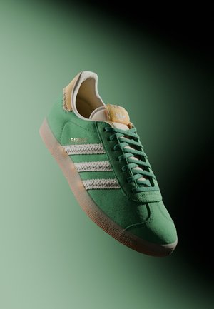 Zapatilla Adidas Gazelle verde con rayas blancas, suela de goma y pestaña amarilla en el talón sobre un fondo degradado verde.