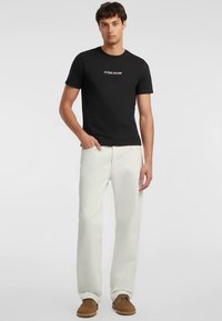 Juoda trumpomis rankovėmis marškinėliai su „Guess Jeans“ logotipu, derinami su baltais tiesaus kirpimo džinsais ir ruda avalyne, stovintys prie paprasto fono.