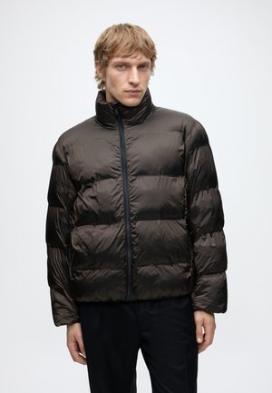 Homme blond portant une veste matelassée noire avec un col haut et une fermeture éclair frontale, debout devant un fond clair uni.