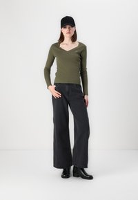Olivenfarget langermet topp med V-hals, kombinert med svarte vide jeans og svarte ankelstøvler. Tilbehøret inkluderer en svart caps.