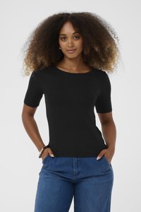 Tânără cu păr ondulat și voluminos, purtând un top scurt cu dungi negre și blugi, cu mâinile în buzunare, pe un fundal neutru.