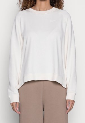 Kobieta w luźnej białej bluzie z długim rękawem w połączeniu z beżowymi spodniami, przedstawiona od ramion do bioder.